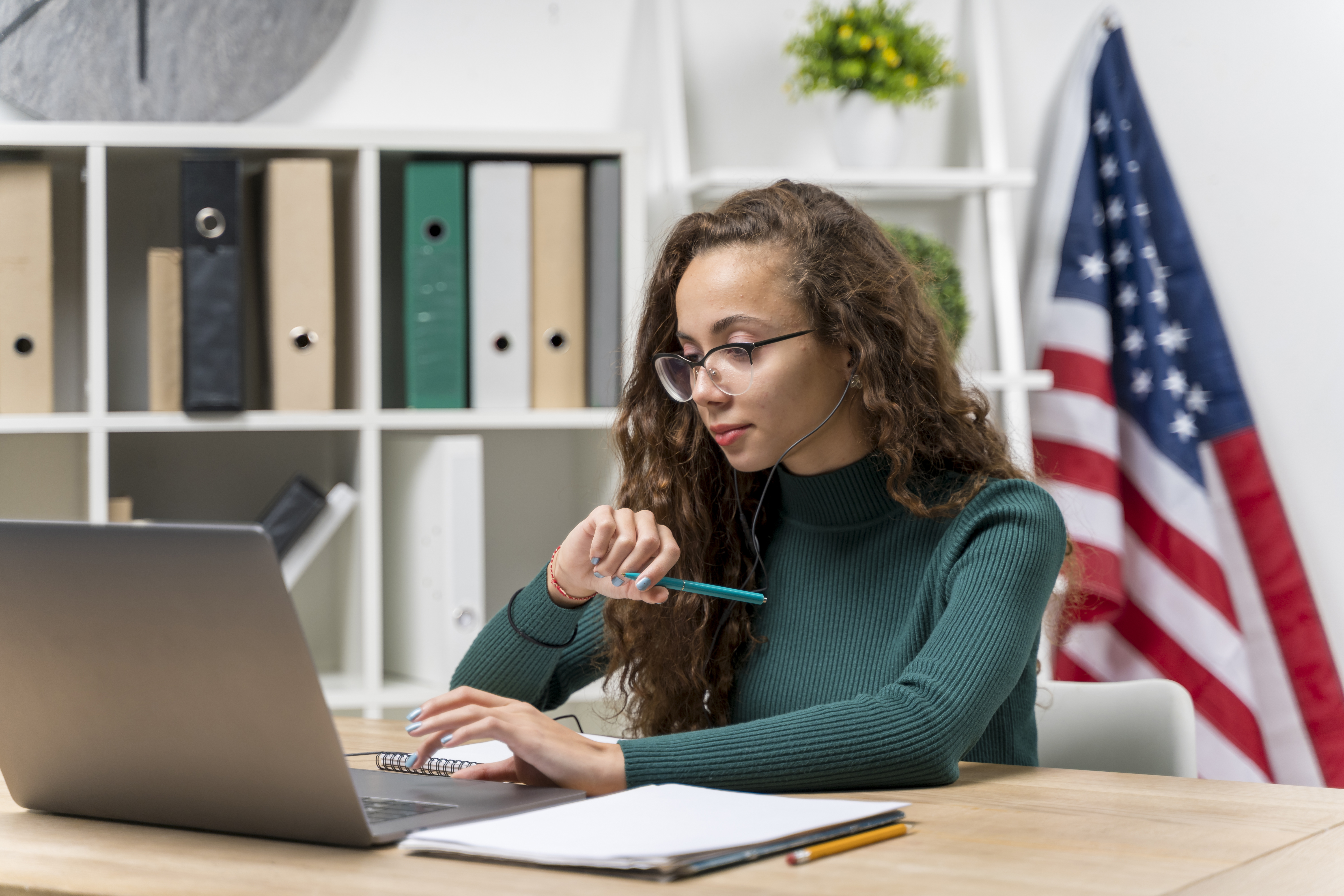 Mujer joven con cabello rizado y gafas, concentrada en su computadora portátil mientras toma notas en un cuaderno, en un entorno de oficina moderno con estanterías y una bandera de Estados Unidos de fondo.