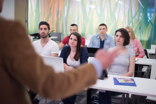 Grupo de estudiantes en un aula moderna, prestando atención a un instructor que está de pie al frente. Algunos estudiantes tienen laptops y tabletas, mientras que el fondo muestra un diseño colorido de hierba.