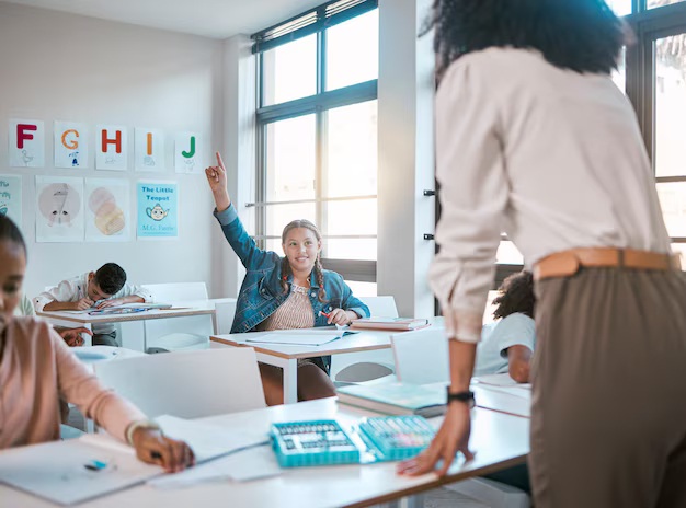 Estudiante levantando la mano en un aula moderna, con una maestra de espaldas y otros alumnos en sus escritorios. Las paredes están decoradas con letras y materiales educativos.