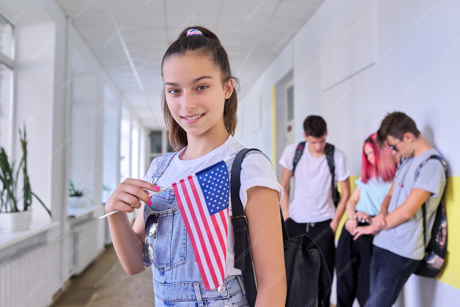 Joven sonriente sosteniendo una bandera de Estados Unidos en un pasillo escolar, con compañeros de fondo. La imagen refleja un ambiente juvenil y de camaradería.
