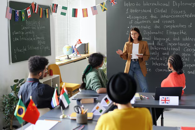 Clase sobre diplomacia internacional con una profesora que presenta a un grupo diverso de estudiantes. La sala tiene banderas de varios países y una pizarra con notas sobre temas globales.