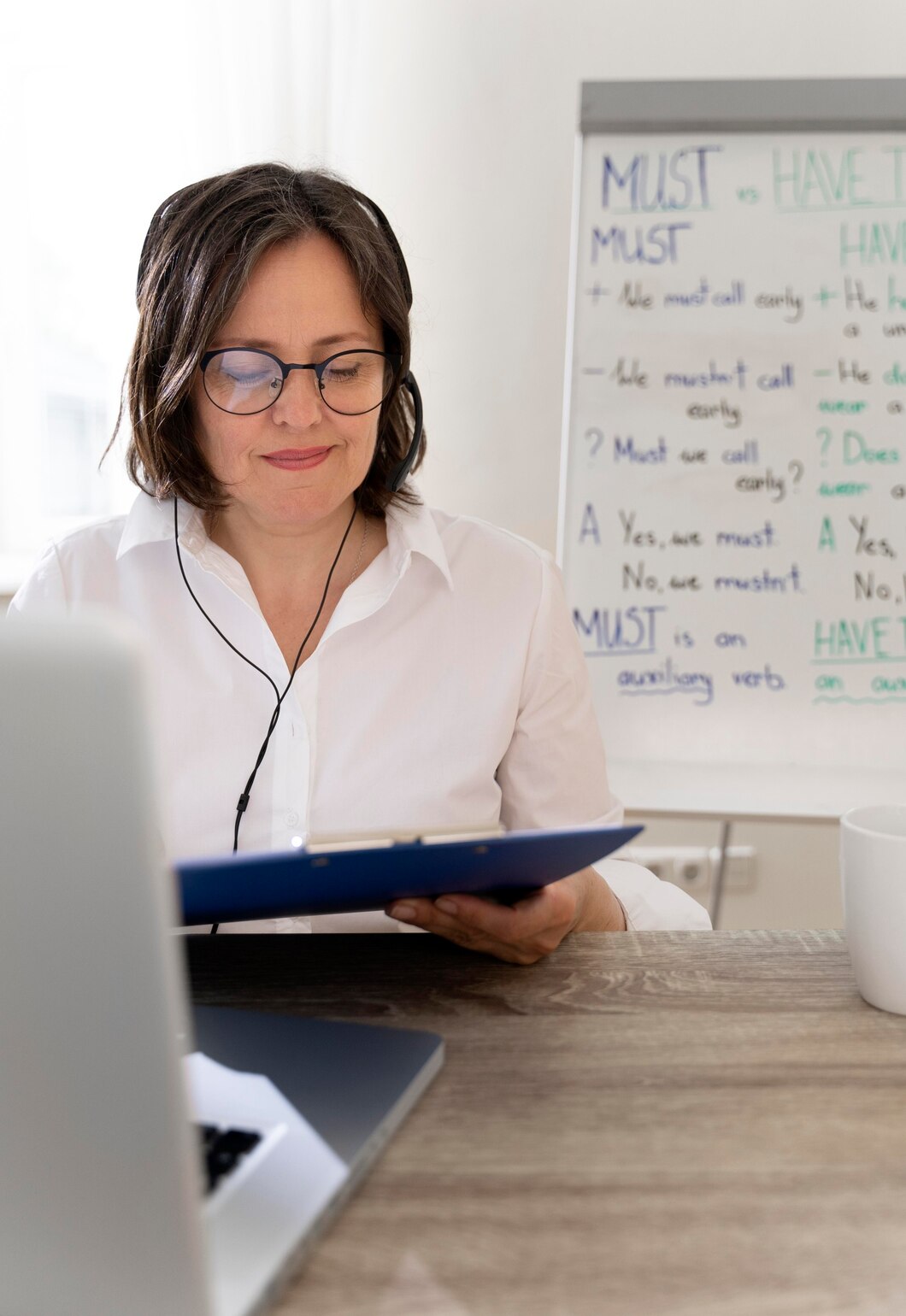 Mujer con auriculares y gafas, revisando documentos en un entorno de trabajo moderno, con una computadora portátil y una pizarra blanca al fondo que muestra ejemplos de gramática en inglés sobre "must" y "have to".