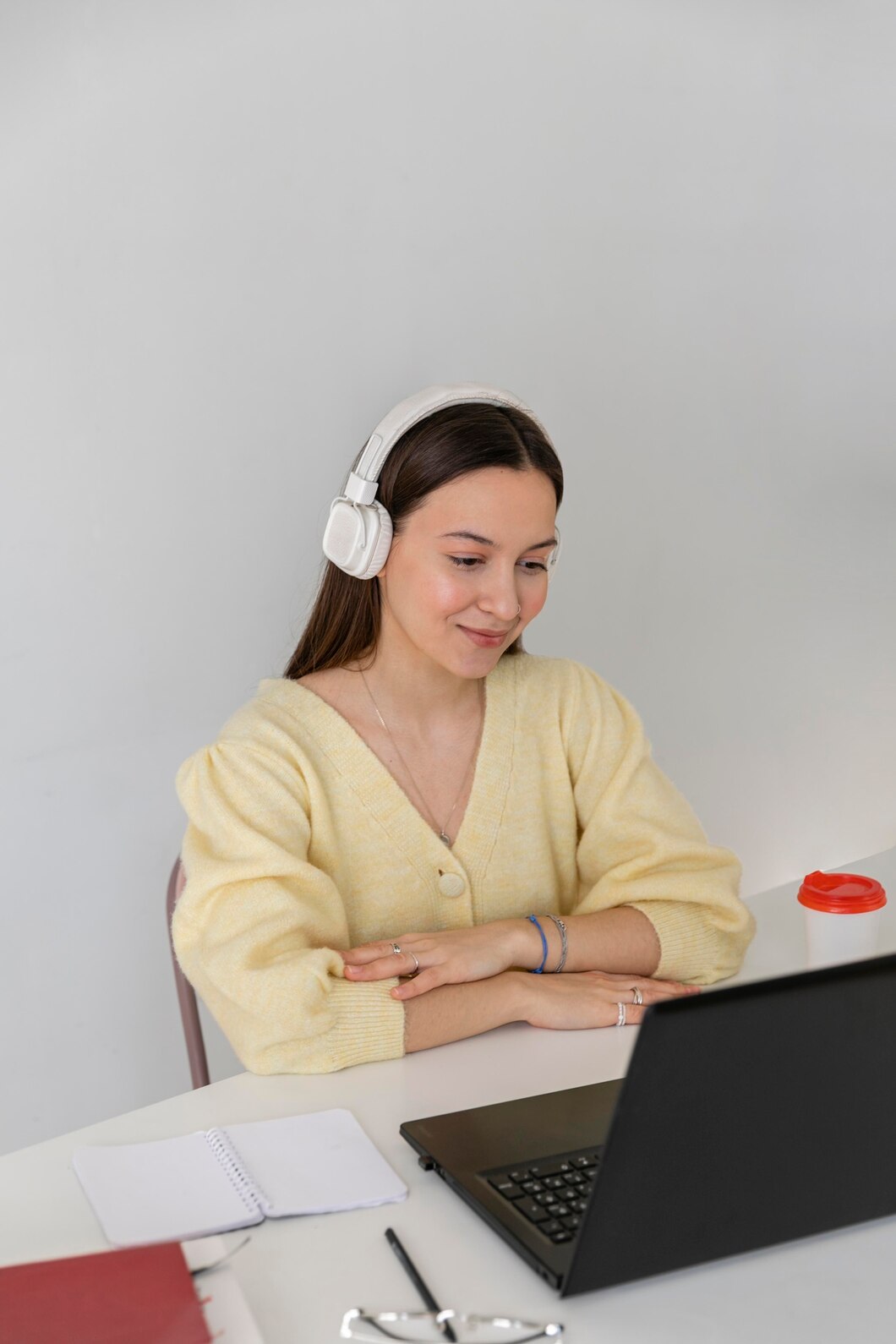 Mujer con auriculares blancos sonriendo mientras trabaja en una computadora portátil negra, rodeada de material de oficina en un ambiente de estudio.