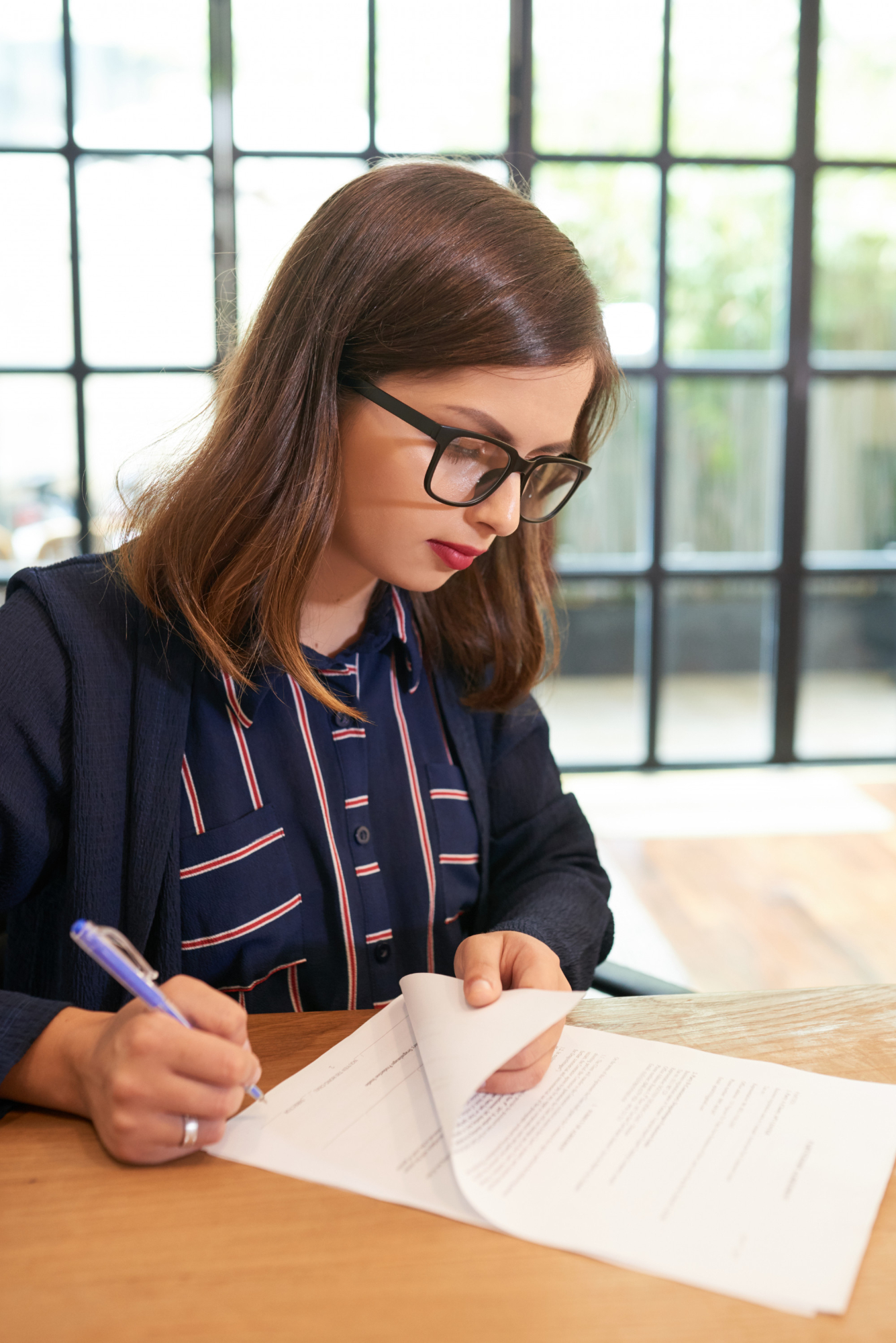 Mujer con gafas escribiendo en un documento en una mesa de madera, en un ambiente luminoso y moderno.
