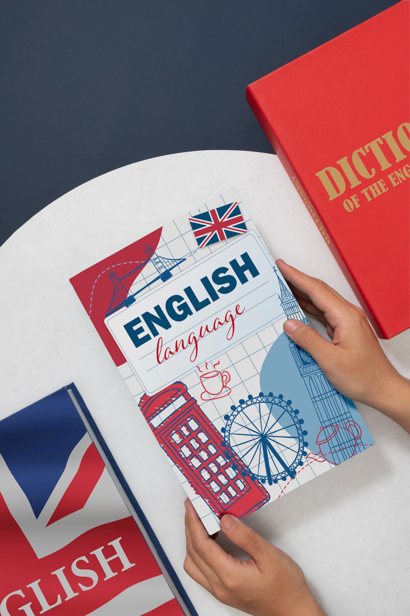 Manos sosteniendo un libro titulado "English Language" con ilustraciones de iconos británicos, junto a un diccionario rojo de inglés y un cuaderno azul con la bandera del Reino Unido, sobre una mesa blanca.