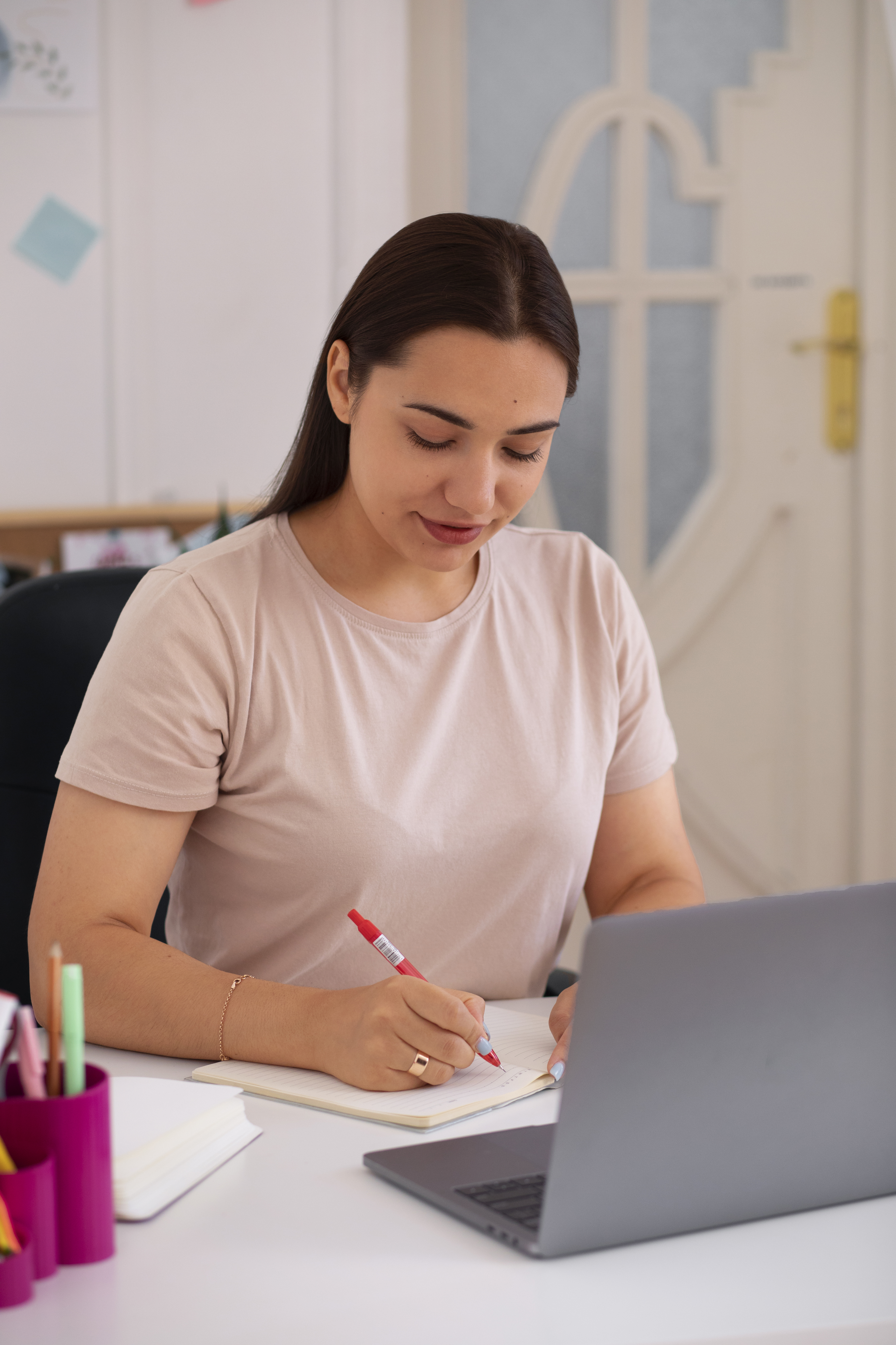 Mujer trabajando en un escritorio moderno, escribiendo en un cuaderno con un marcador rojo, mientras utiliza una laptop gris. El ambiente es luminoso y ordenado, con elementos de papelería coloridos.